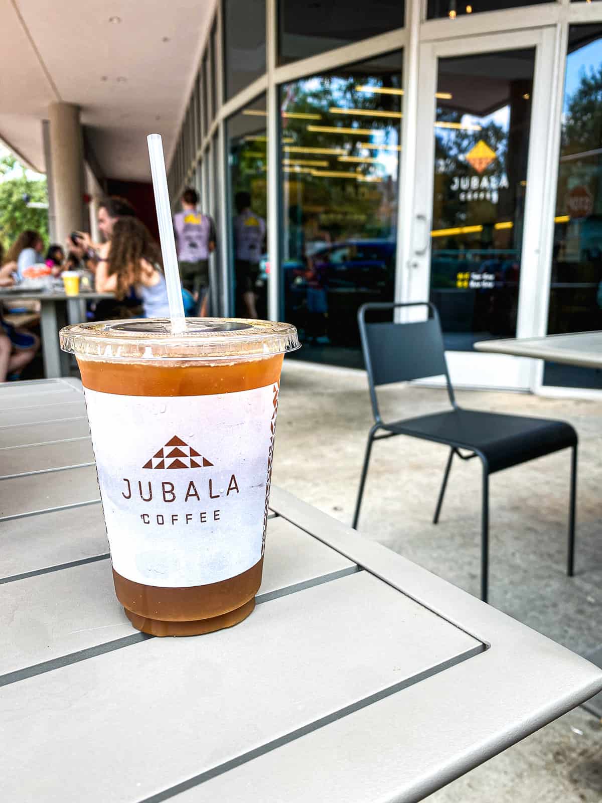 An iced coffee in a plastic cup that says Jubala on it on a table outside with large windows