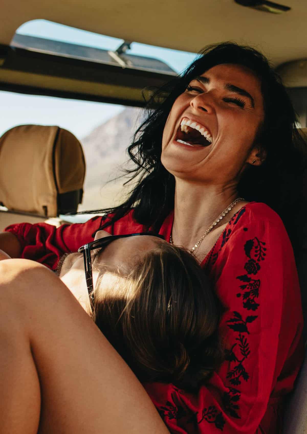 A woman sitting in the car laughing and her husband's head is on her lap. 