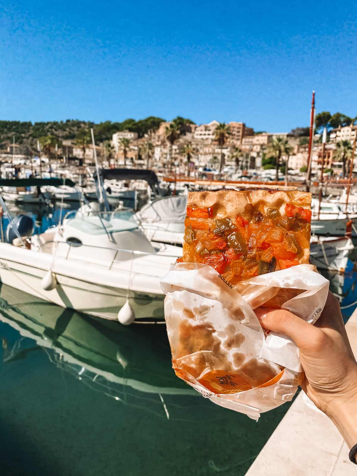 A hand holding bread with vegetables on it by a harbor with boats