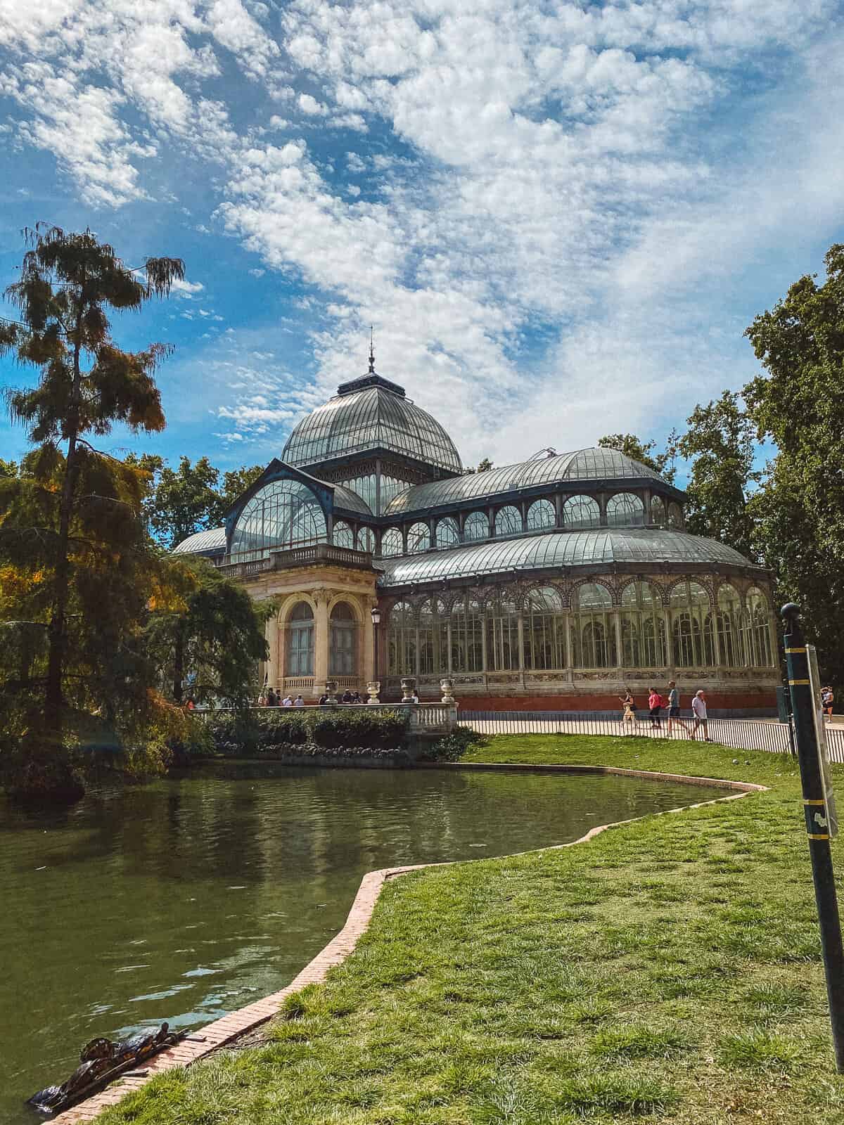 The Crystal Palace in Madrid's Retiro Park, a striking glass and metal structure reflecting the sunlight, set beside a tranquil pond with lush greenery and visitors enjoying the serene surroundings