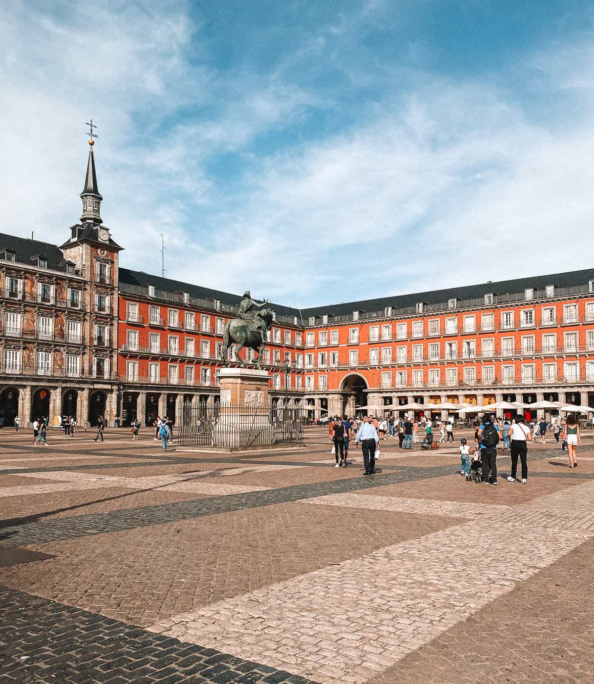 A lively view of Plaza Mayor in Madrid showcasing the statue of King Philip III at the center, with visitors walking around the spacious cobblestone square surrounded by classic red facades and arches.