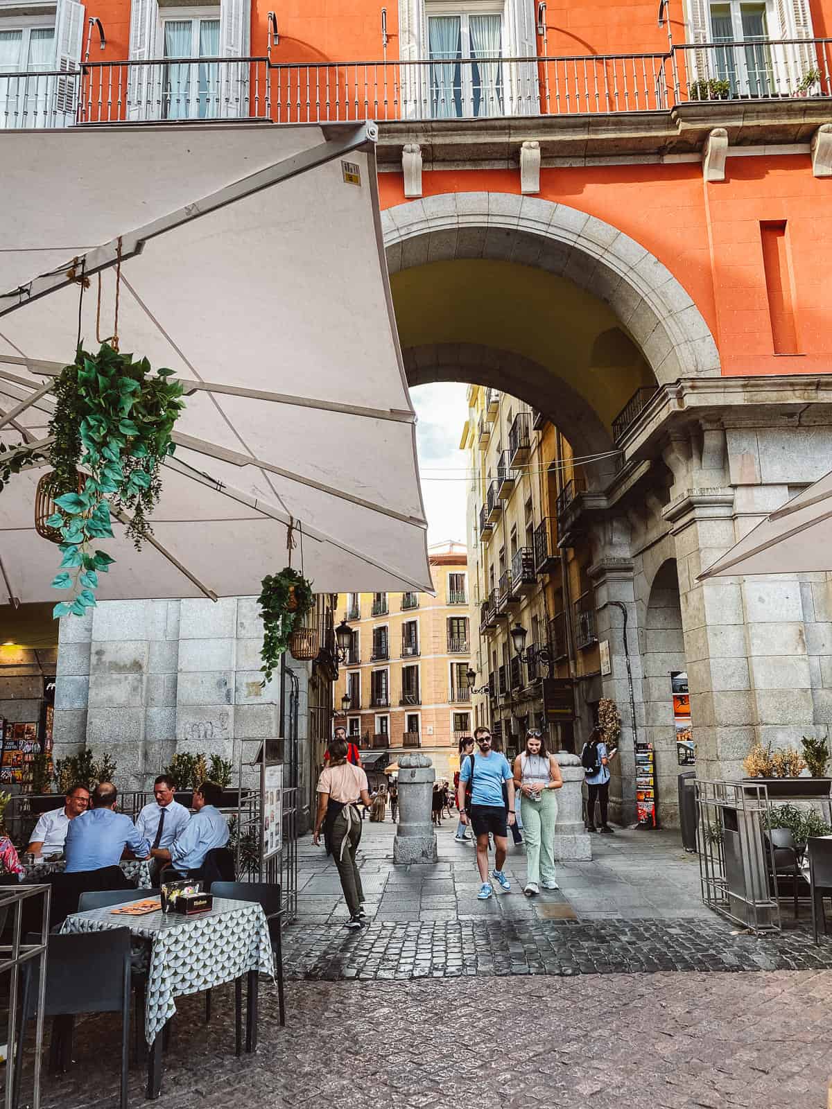 A vibrant street scene in Madrid with people walking under the arches of Plaza Mayor, outdoor seating at a café where patrons are dining, and traditional Spanish buildings with balconies overlooking the square.