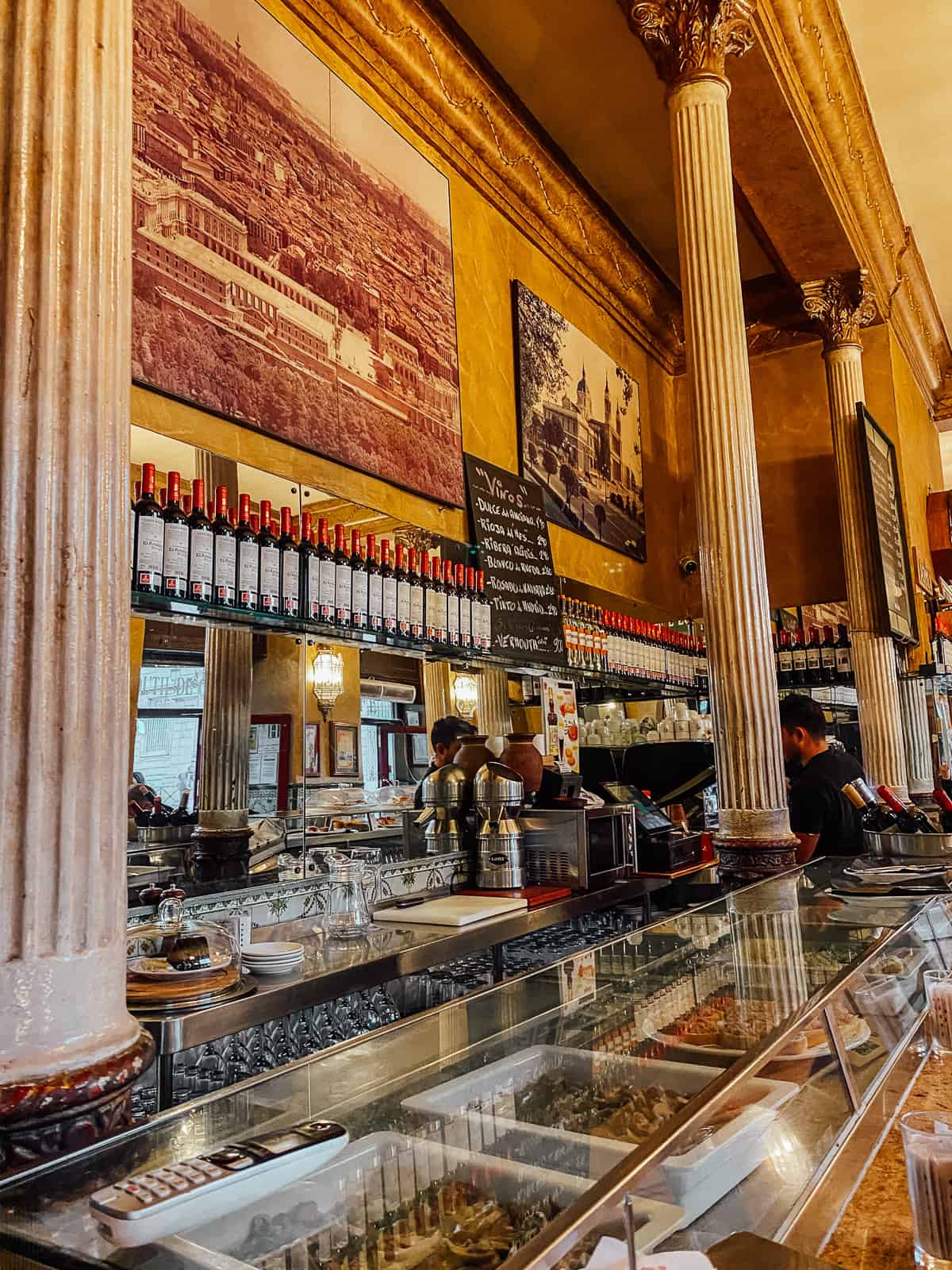 Interior of a traditional Madrid café with tall Corinthian columns and a long bar displaying rows of wine bottles. The walls are adorned with sepia-toned photographs of historical landmarks, and baristas are attending to customers in the background