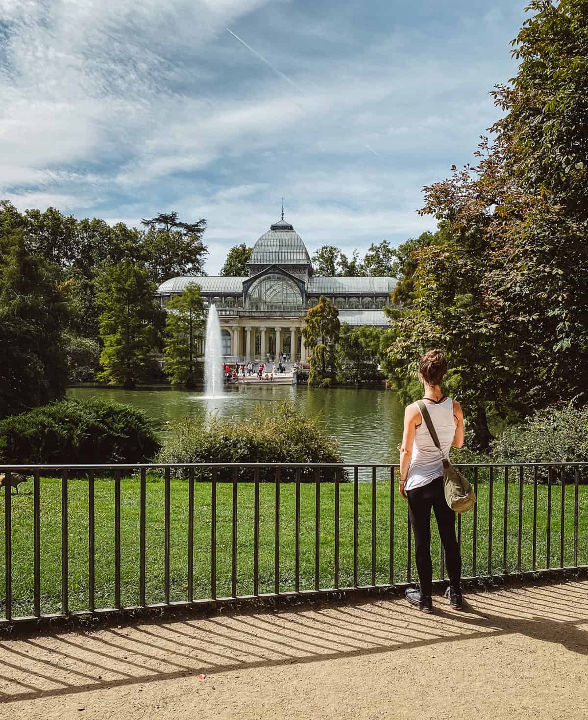a woman standing in a green park in front of a fountain and the crystal palace in Madrid