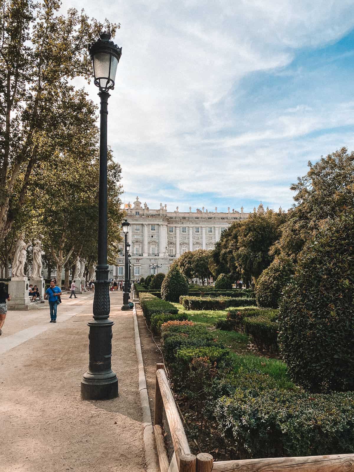 A classic street lamp illuminates a path in Retiro Park, with the Royal Palace of Madrid, Spain, visible in the distance, showcasing the historical and cultural landmarks of the city