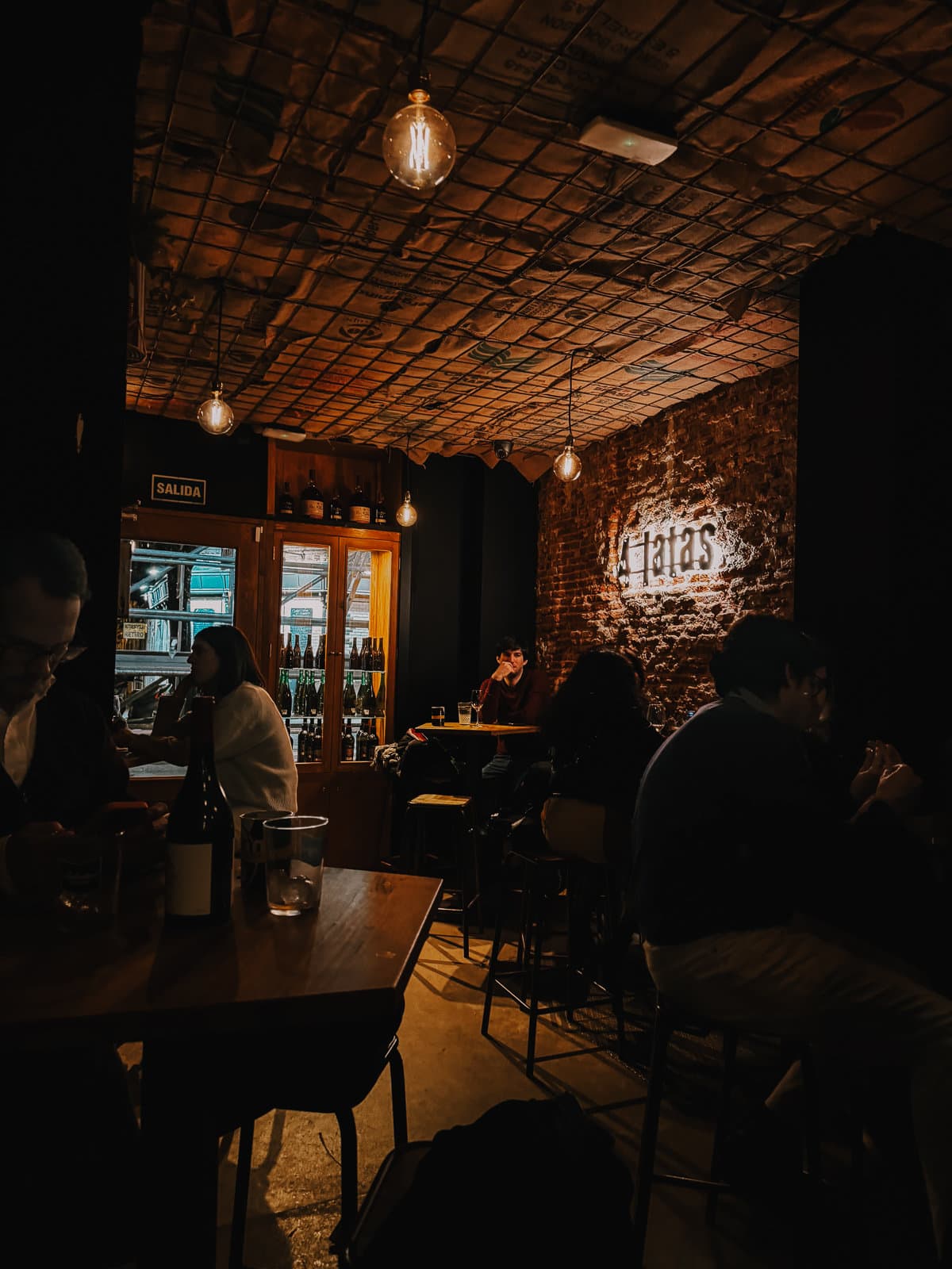 Dimly lit cozy tapas bar with exposed brick walls and warm lighting, patrons dining and conversing in a relaxed atmosphere, highlighting the word 'tapas' illuminated on the wall