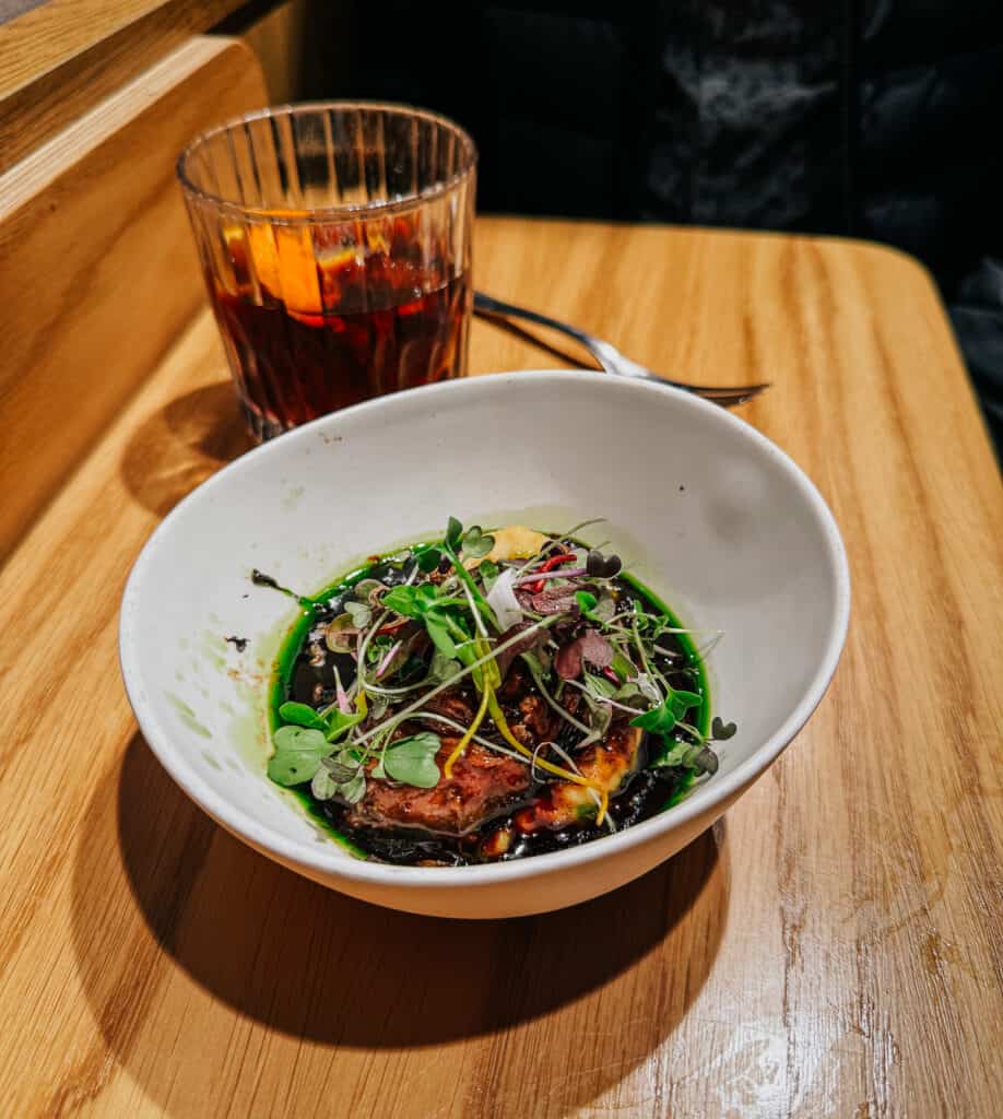 Gourmet dish served in a white bowl on a wooden table, featuring colorful microgreens and a drizzle of balsamic glaze over a pork cheek, showcasing a vibrant and appetizing presentation.
