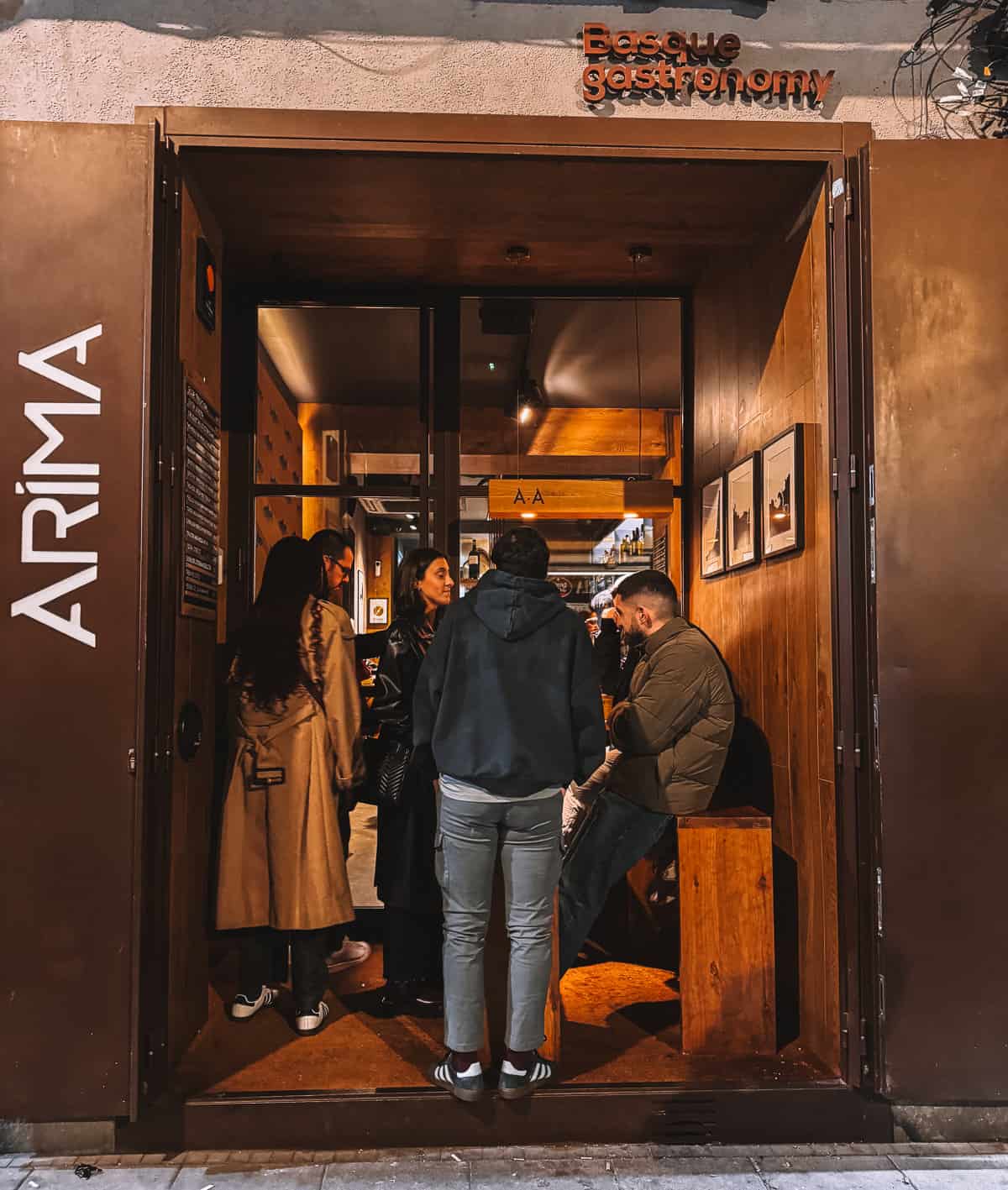 Nighttime scene outside 'ARIMA Basque Gastronomy' restaurant entrance, with people engaging and waiting, emphasizing the lively urban dining experience.