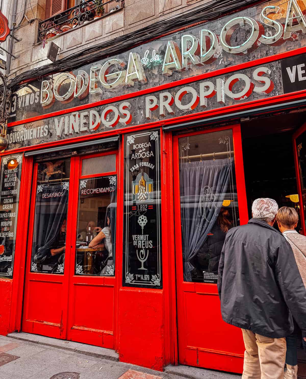 Exterior view of Bodega Ardosa, a vibrant red facade with large windows, patrons visible inside, and bold signage promoting wines and vermouths
