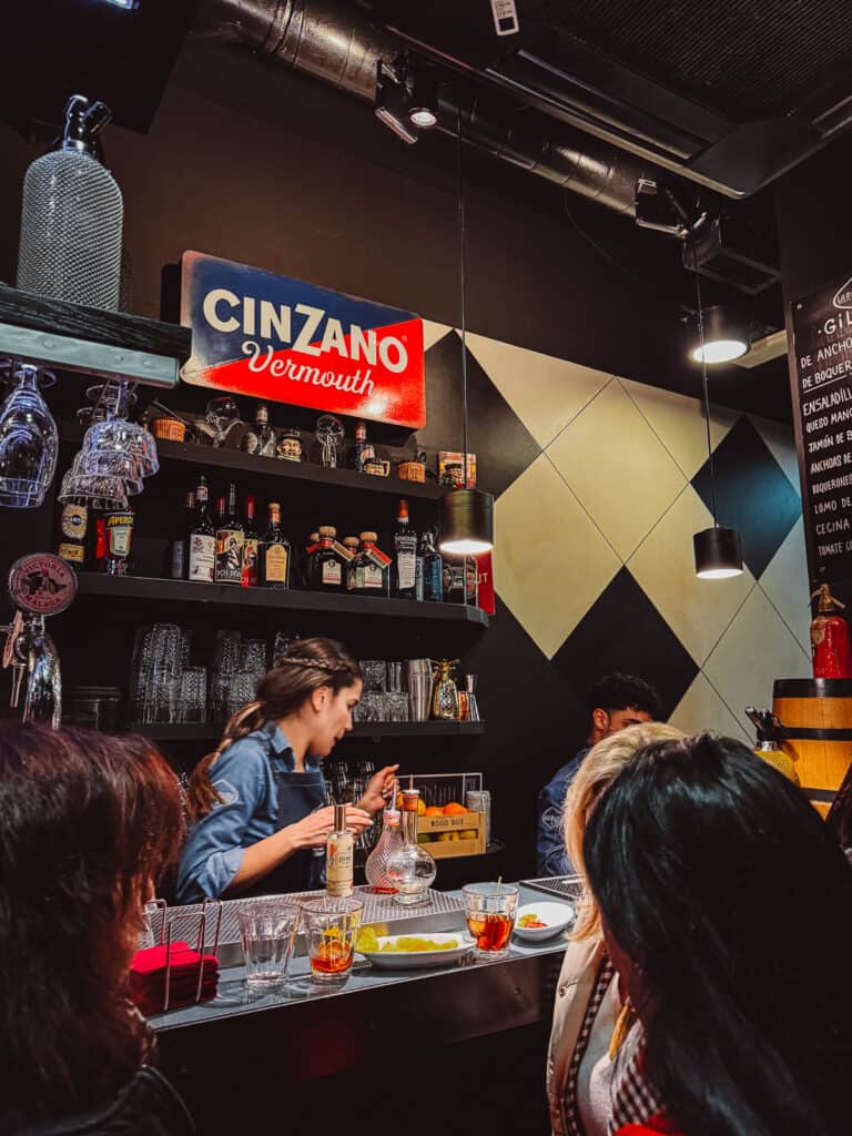 Patrons enjoying drinks at a bar with a prominent 'CINZANO Vermouth' sign, various spirit bottles on the shelves, and a bartender in motion