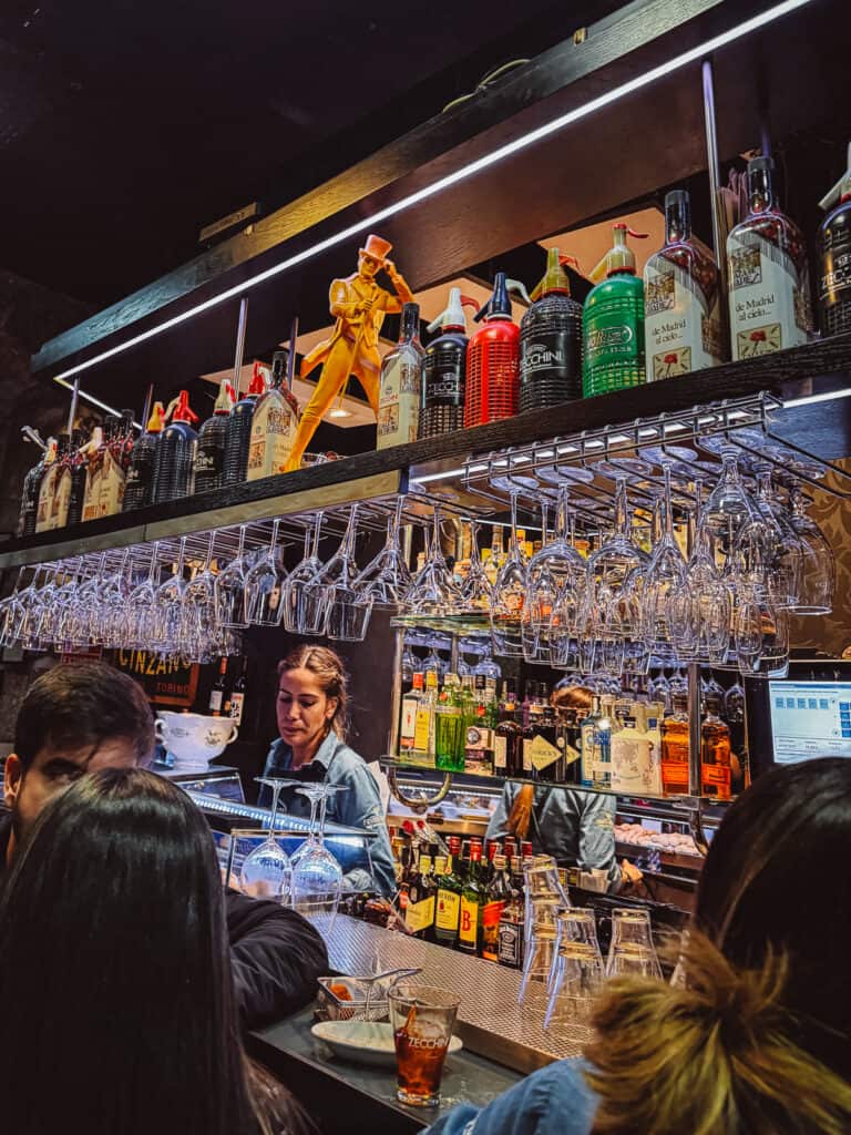 Busy bar atmosphere viewed from the customer side, showing a bartender serving drinks with an array of spirit bottles in the background, and a vibrant sculpture of a man in a yellow suit atop the shelf