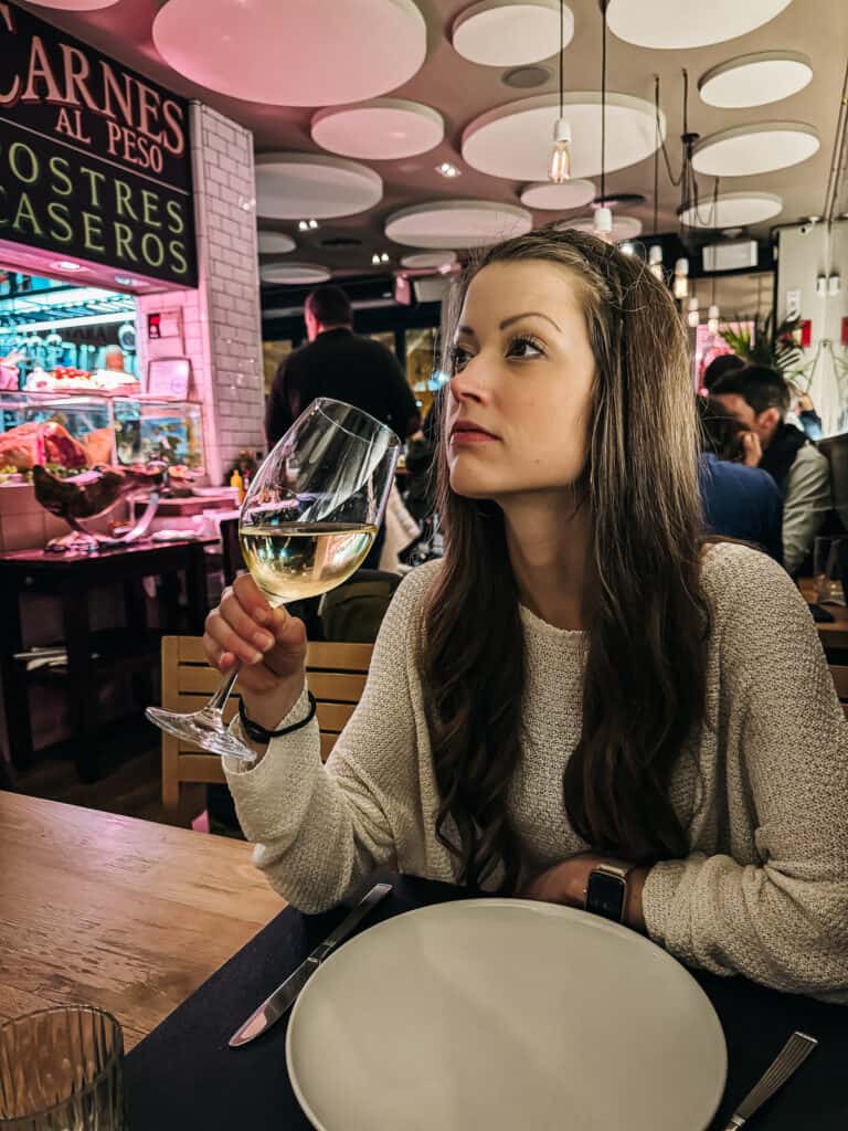 A woman in a cozy sweater savoring a glass of white wine, with a focus on her thoughtful expression, in a bustling restaurant with a display counter in the background