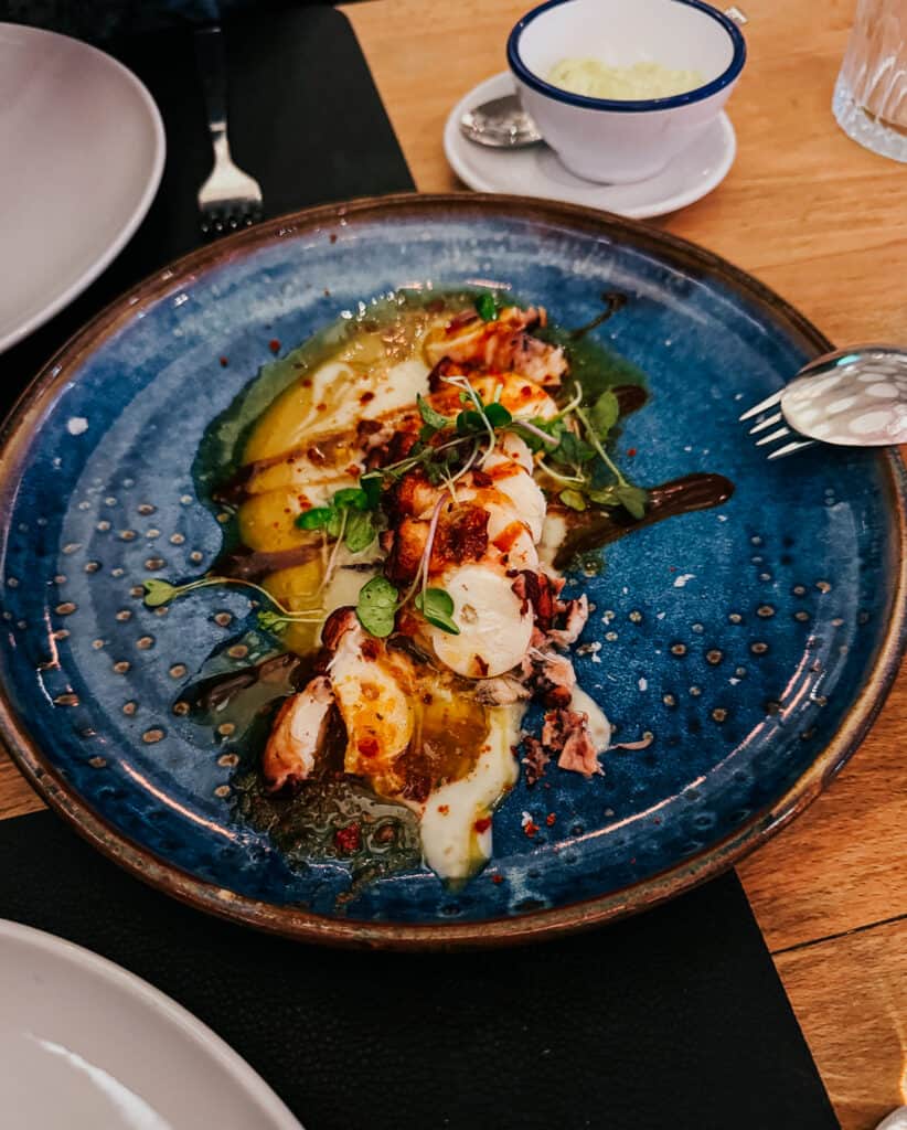 A blue ceramic plate holds an artistically arranged octopus dish with sliced potatoes, sprinkled with herbs and spices, served next to a small cup of aioli, depicting a fine dining experience