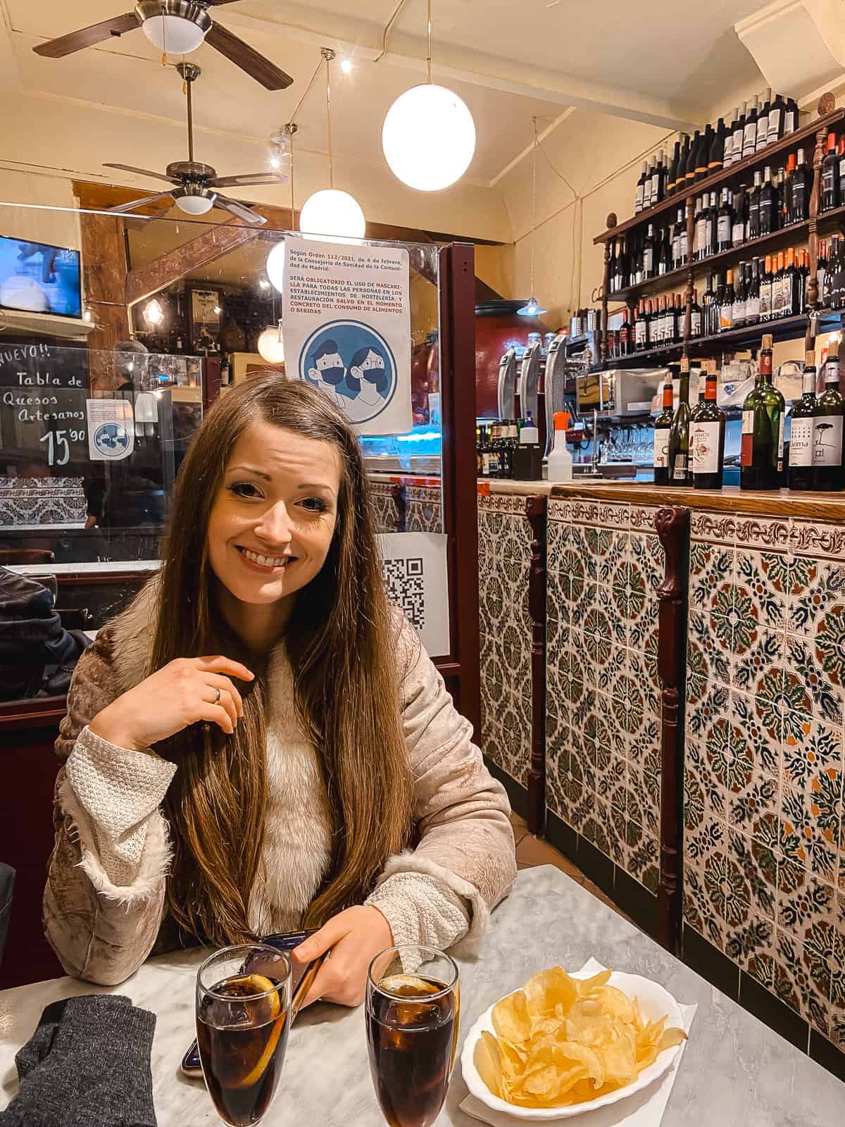 A smiling woman seated at a bar with traditional tiles, enjoying a glass of dark beverage with a bowl of chips, creating a cozy dining atmosphere.
