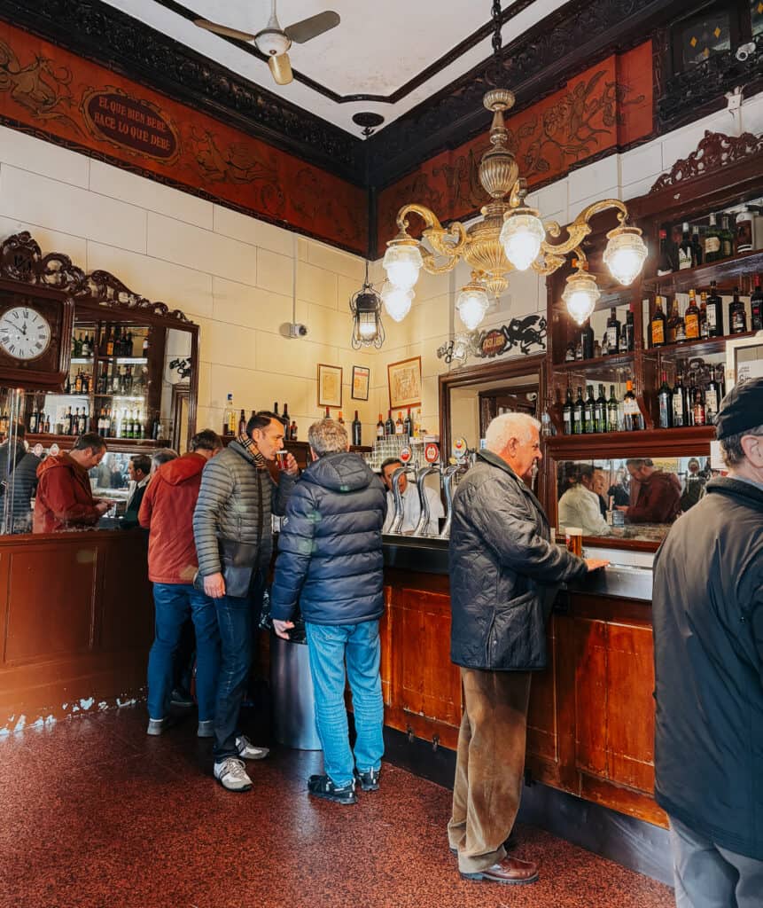 Interior of a traditional bar with patrons at the counter, ornate chandeliers, and antique decor, illustrating a classic European pub ambiance