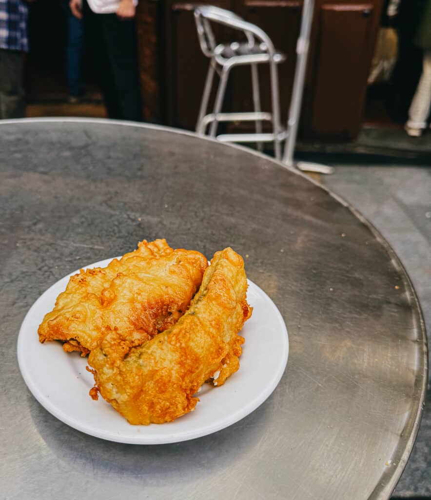 A close-up of a plate with two pieces of crispy battered fish on an outdoor metal table, symbolizing a casual street food experience
