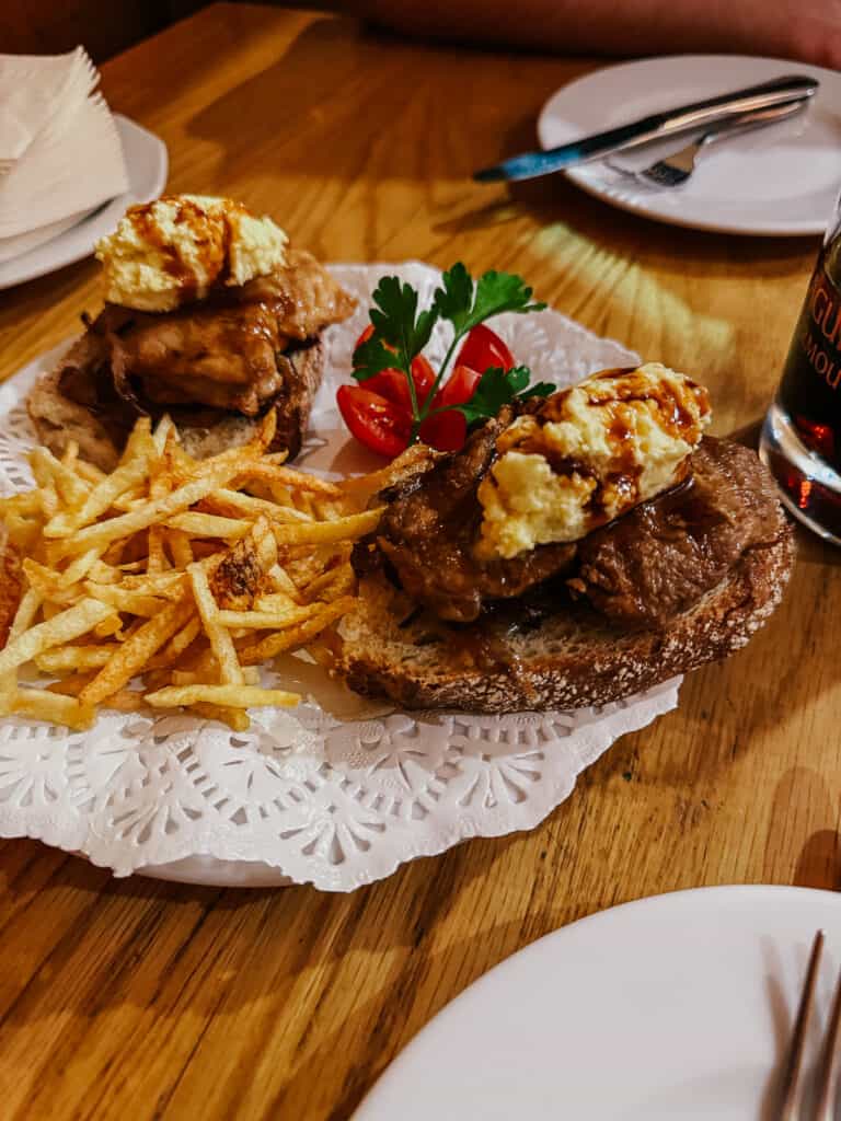 A close-up of a hearty meal featuring an open-faced chicken sandwich topped with scrambled eggs, served with a side of shoestring fries, garnished with parsley and a cherry tomato