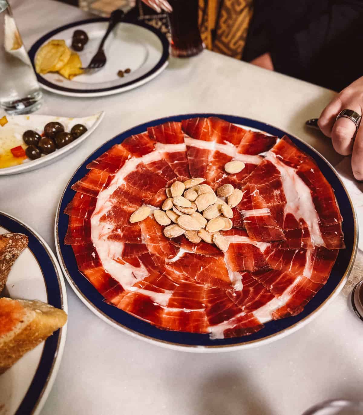 A large, round plate filled with thinly sliced jamón ibérico garnished with a pile of almonds in the center, partially surrounded by other tapas dishes, showcasing a vibrant dining setting.