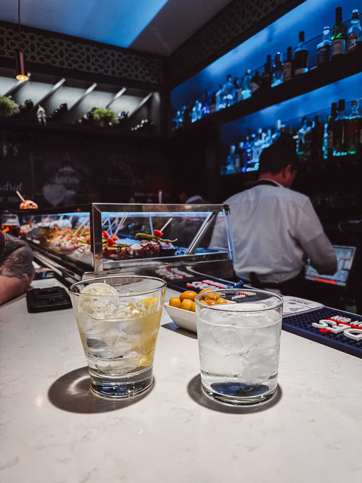 Two cocktails on a marble bar counter with a vibrant tapas display in the background, showcasing the lively atmosphere of a Spanish bar.