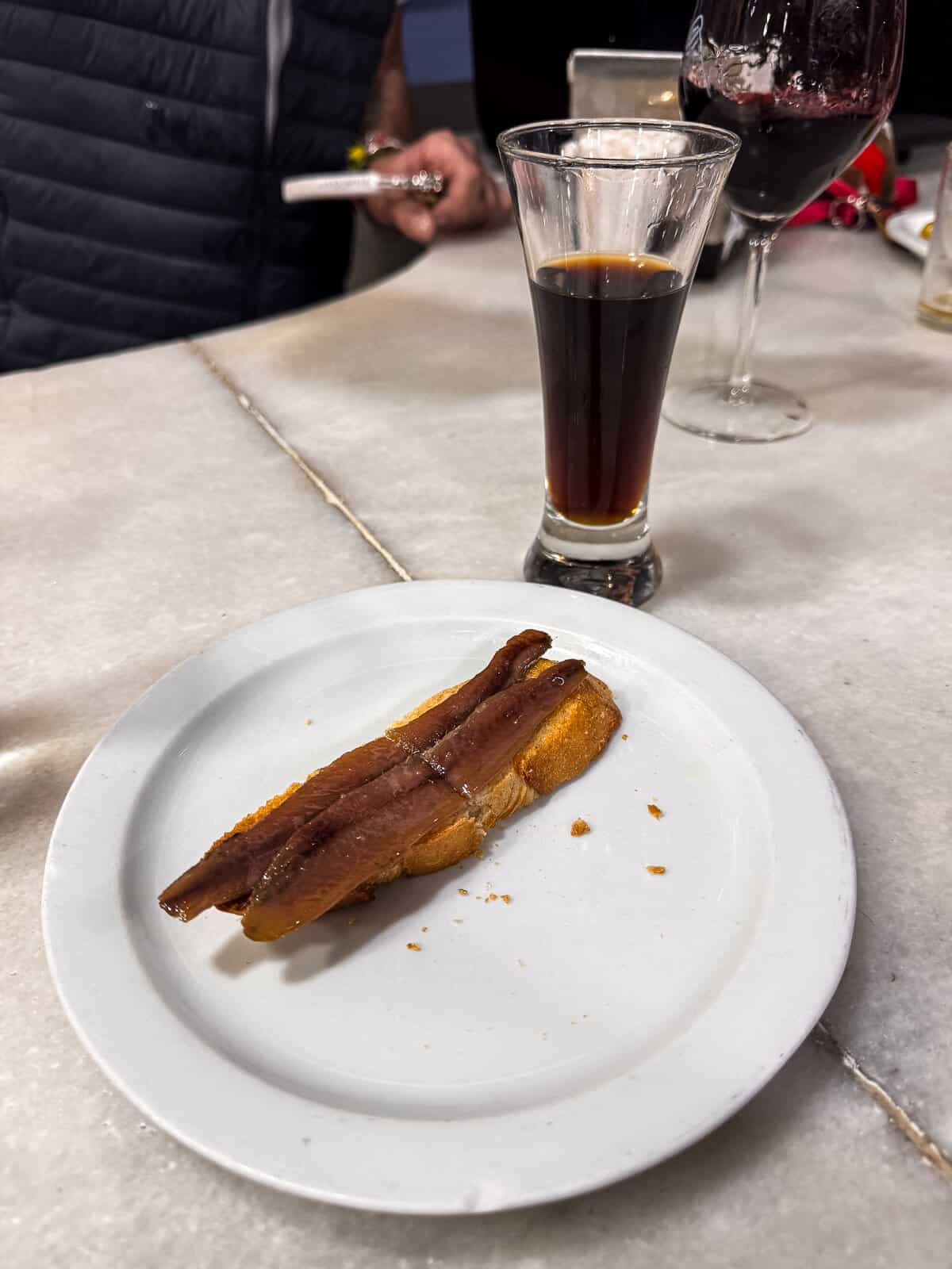 A close-up of a simple Spanish snack on a white plate, featuring crispy bread topped with anchovies, beside a tall glass of dark vermouth, set on a marble table with casual dining background.