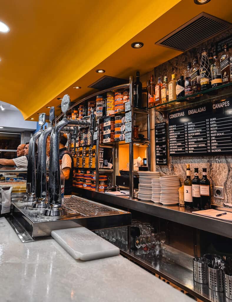 Vibrant and busy Spanish bar interior with a yellow ceiling, featuring a variety of canned goods displayed behind the bar and a detailed menu board, creating an authentic tapas bar atmosphere.