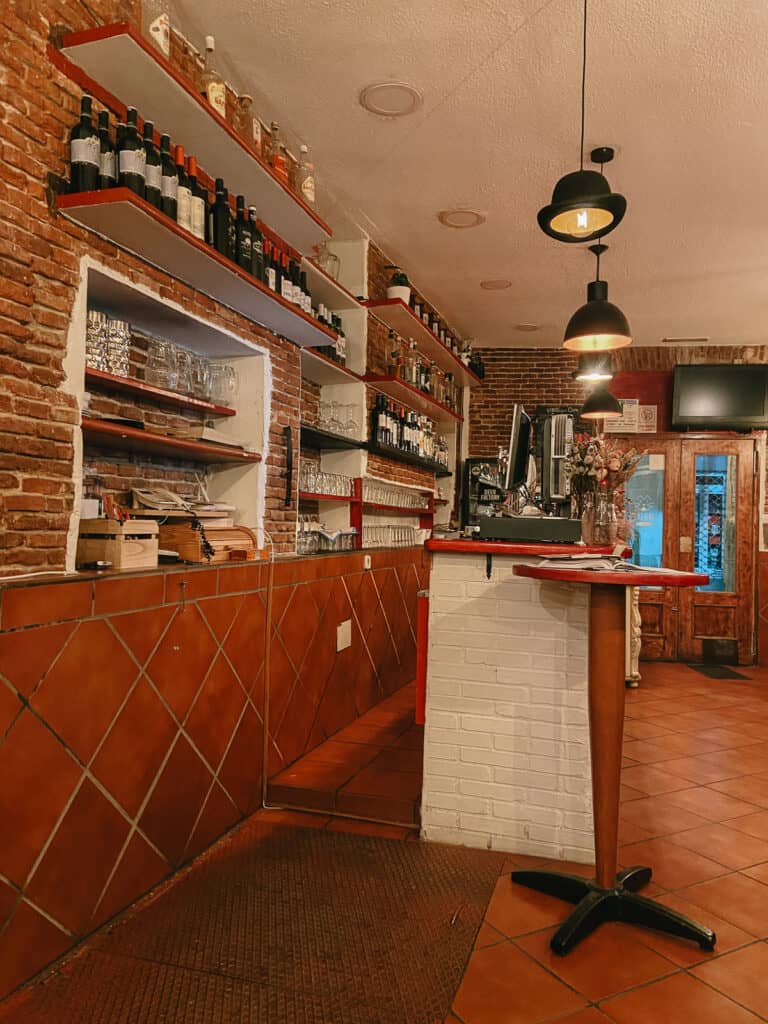Rustic bar interior with exposed brick, shelves stocked with wine bottles, and a warm, inviting atmosphere for a casual drink.