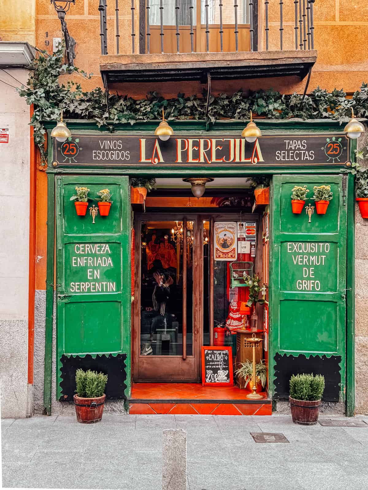 The vibrant green exterior of La Perejila bar in Madrid, adorned with flower pots and a sign advertising selected wines and tapas, invites passersby into its warm, welcoming atmosphere.