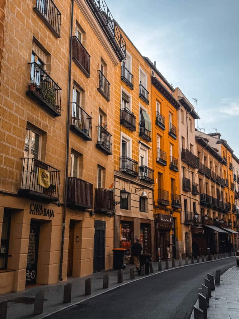 A tranquil scene of Cava Baja street in Madrid, lined with historic buildings painted in warm hues, with balconies overlooking the quiet, cobblestoned street in the early evening.