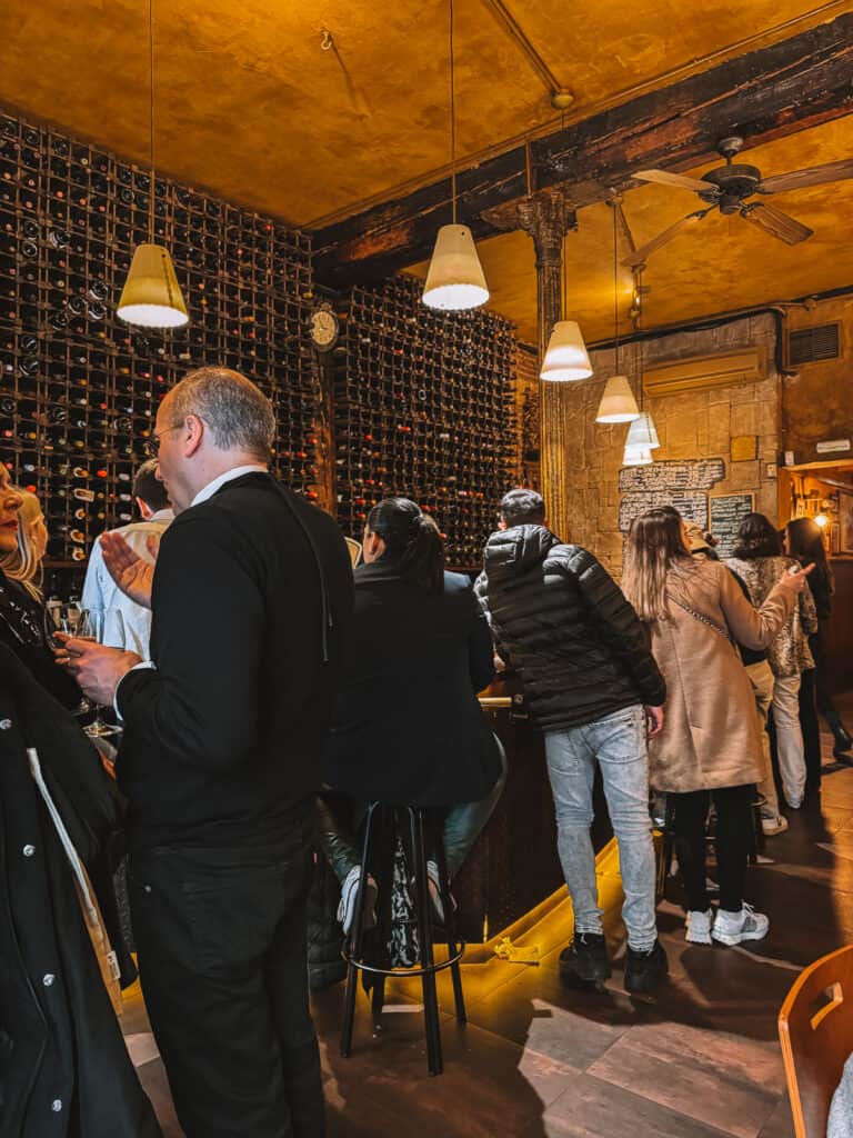 Patrons enjoy drinks and conversation in a bustling Madrid wine bar, surrounded by walls filled with an extensive array of wine bottles, creating an atmosphere of relaxed socializing and wine appreciation