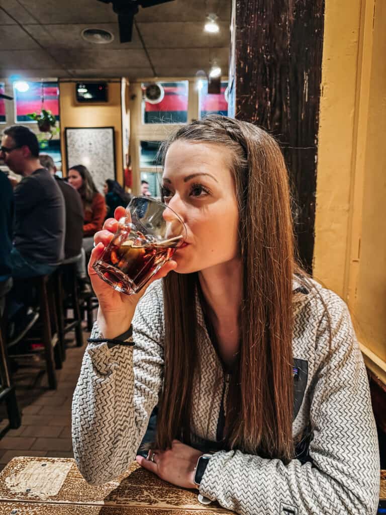 A woman in a patterned sweater sipping from a glass of vermouth in a cozy bar setting, with a soft focus on the bustling background
