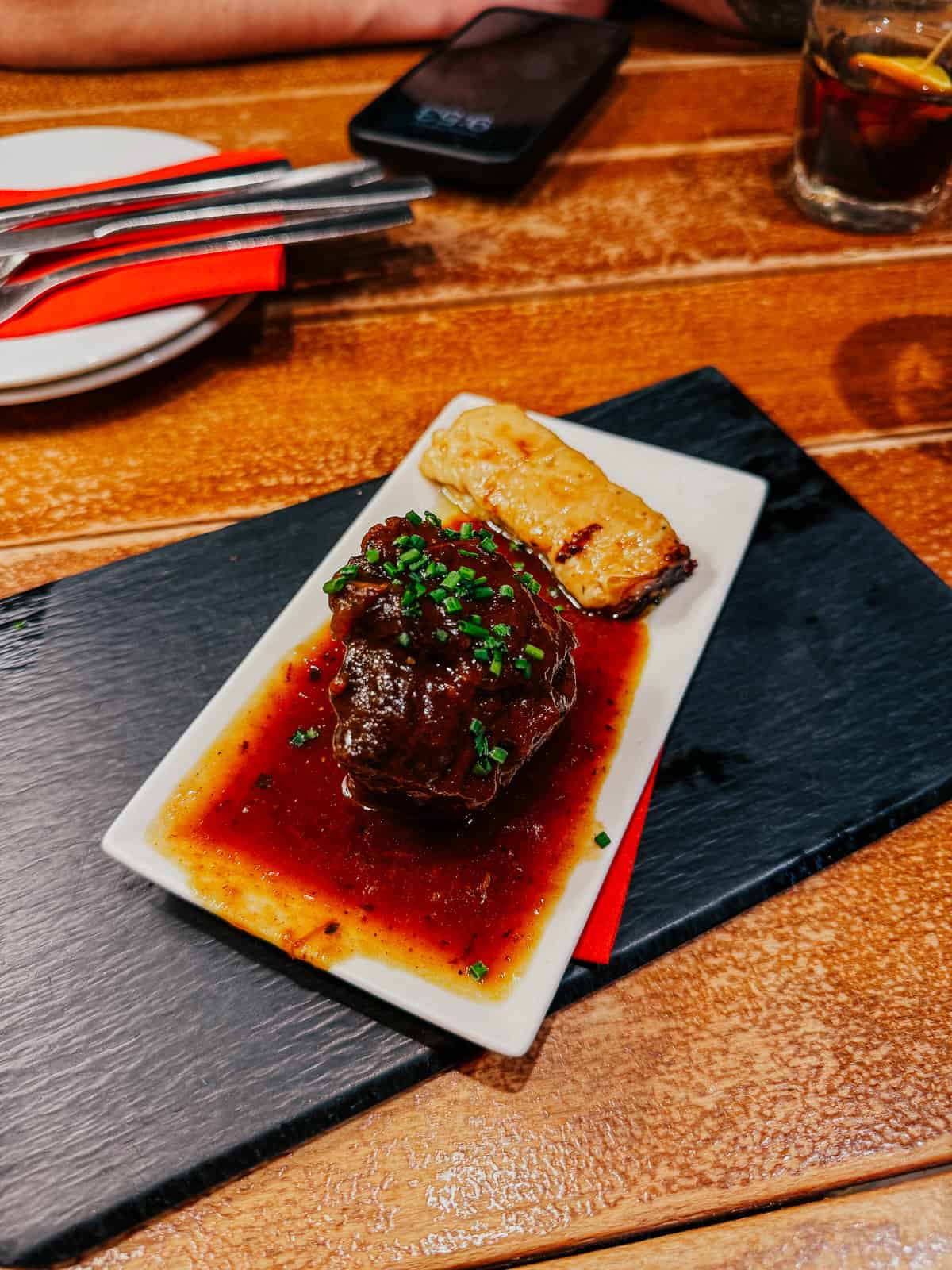 A succulent piece of braised meat in a rich brown sauce, paired with a cheesy gratin, served on a rectangular white plate over a slate board.