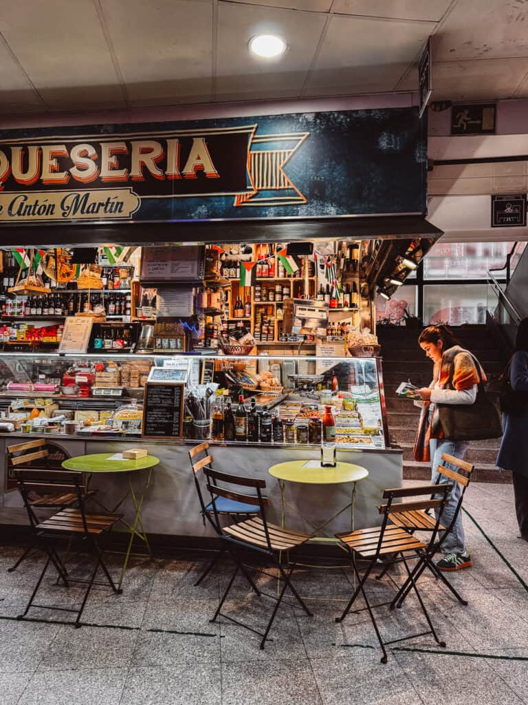 A cozy cheese shop at Mercado de Antón Martín in Madrid, with its distinctive blue sign, showcases a diverse selection of cheeses and gourmet products, inviting market-goers to explore Spanish flavors