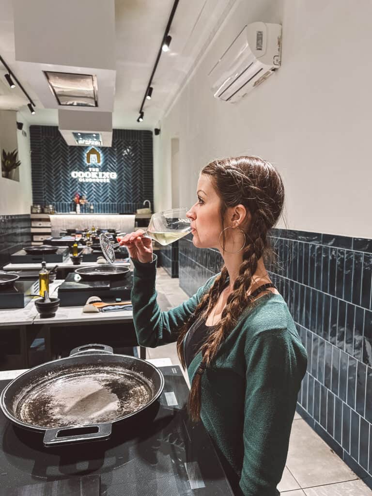 A woman savoring a glass of white wine in a modern cooking school in Madrid, with a clean, sleek kitchen setting that hints at an intimate culinary experience