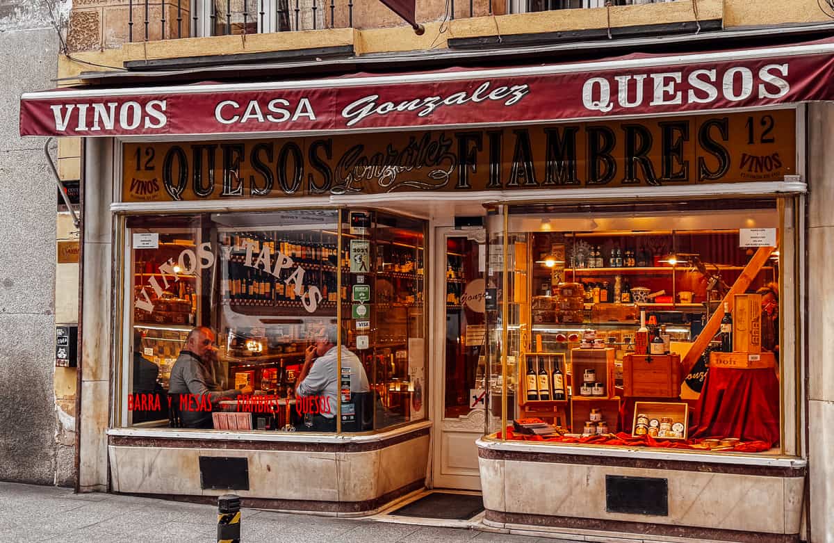 The traditional façade of Casa Gonzalez in Madrid, featuring a red awning, displays a selection of wines and cheeses, with two men engaged in conversation by the storefront, reflecting the city's authentic culinary culture.