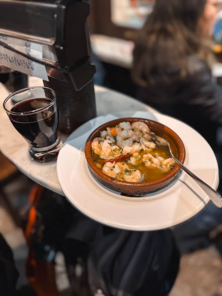 A close-up view of a traditional Spanish meal, featuring a terracotta bowl of garlic shrimp, alongside a glass of red wine, served on a bar counter, evoking the cozy atmosphere of a Madrid tapas bar.