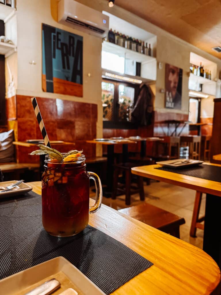 A refreshing sangria in a mason jar, garnished with a citrus twist, sits on a wooden table in a warmly lit, modern Madrid restaurant, inviting a taste of local Spanish refreshments.