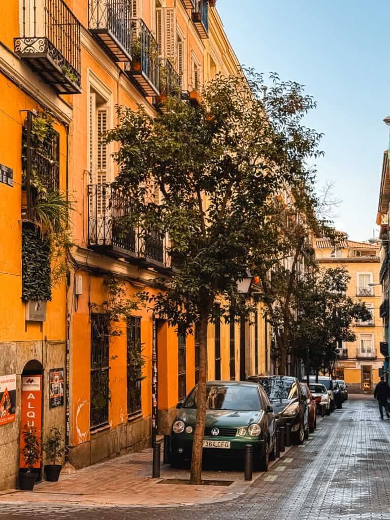 A picturesque street in Madrid with orange and yellow facades, balconies adorned with plants, and cars parked alongside, exuding the warmth of a sunny Spanish day.