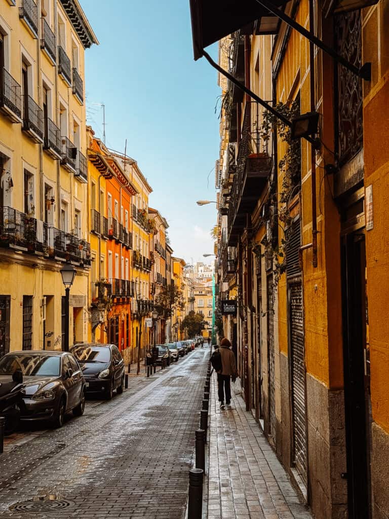A narrow cobblestone street in Madrid lined with colorful buildings and balconies, with a solitary figure walking in the distance, capturing the quaint charm of the city.