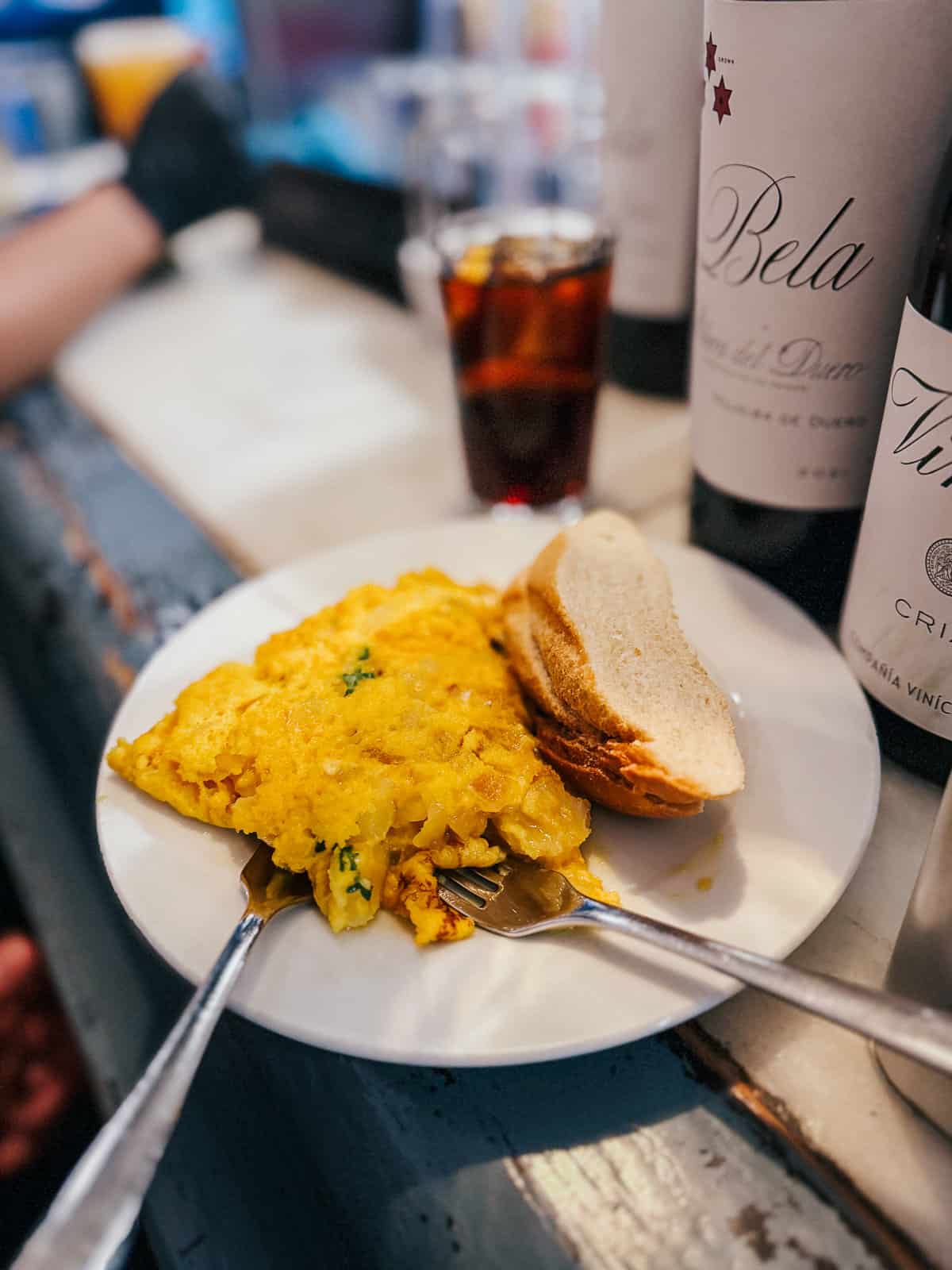 Traditional Spanish omelette served with bread on a white plate, accompanied by a glass of vermouth, set against a backdrop of a busy bar