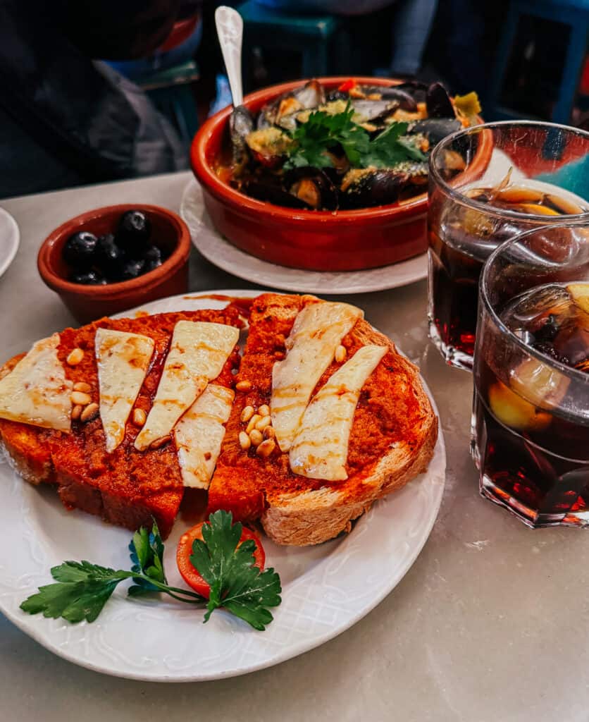 A Spanish culinary delight of tomato bread topped with melted cheese and pine nuts, a bowl of mussels, and a glass of vermouth on a marble tabletop.