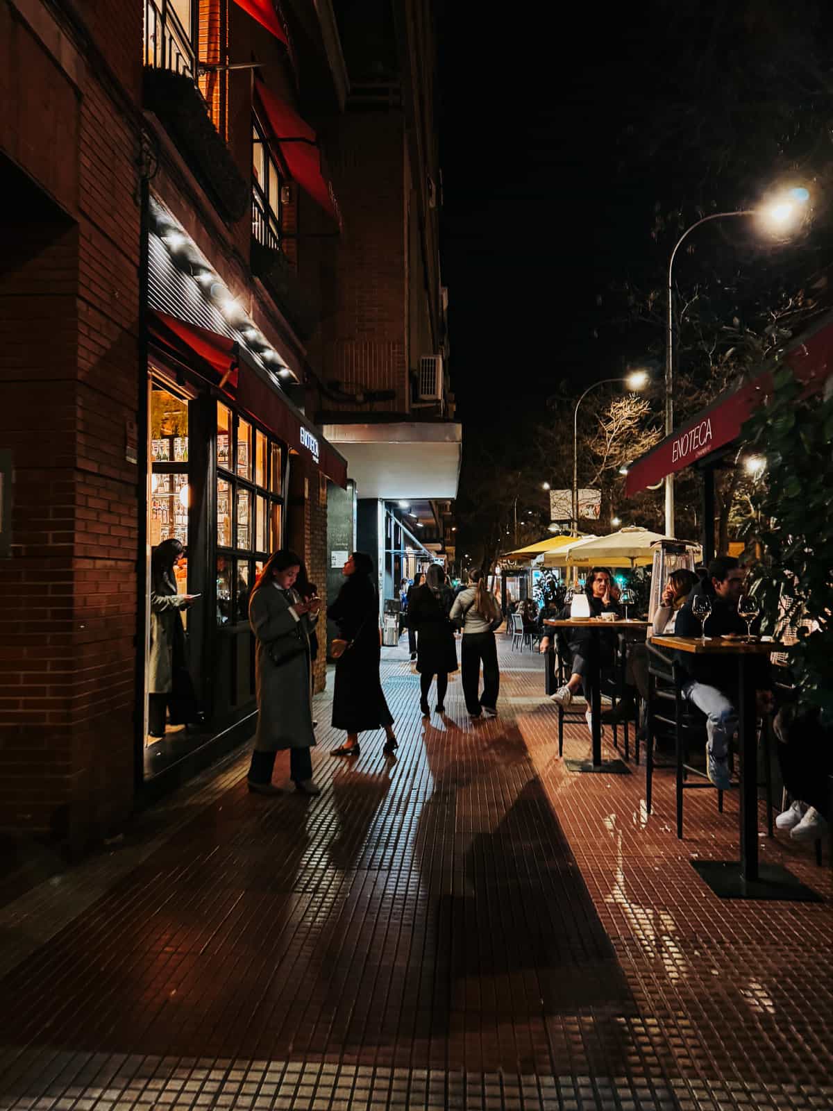 Evening view of a lively street scene outside a row of restaurants and bars, with people walking and dining alfresco, illuminated by street lamps and warm indoor lighting