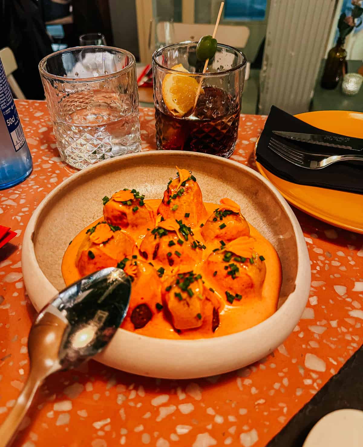 A dish of patatas bravas dressed in a creamy sauce on a terrazzo table, accompanied by a glass of water and cocktail, epitomizing a modern tapas experience.