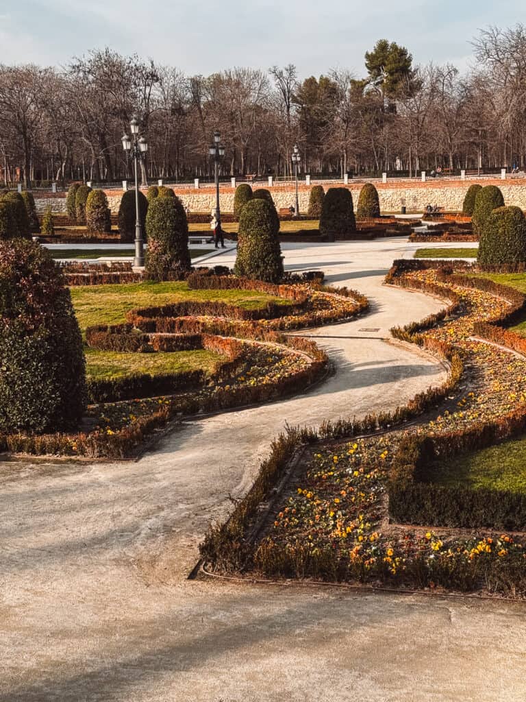 The carefully manicured hedges and paths of the Retiro Park in Madrid present a serene, geometric arrangement, signaling the onset of autumn with scattered golden leaves.