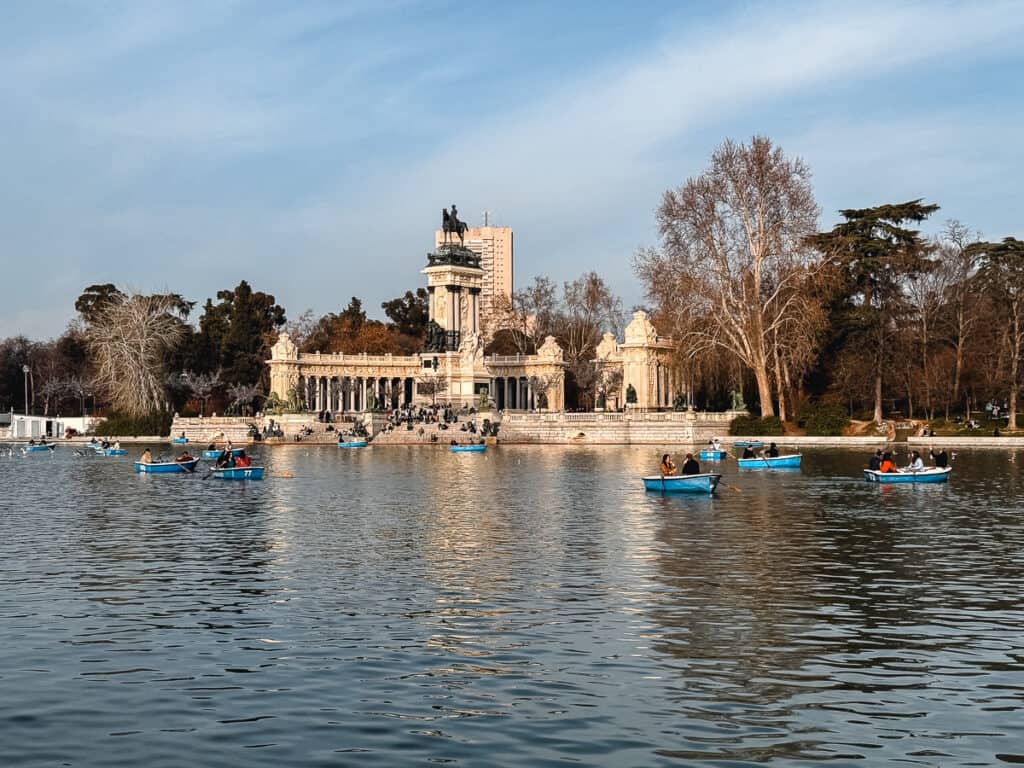 Rowboats float gently on the calm waters of the large pond in Madrid's Retiro Park, with the ornate Monument to Alfonso XII and visitors in the background under a clear sky.
