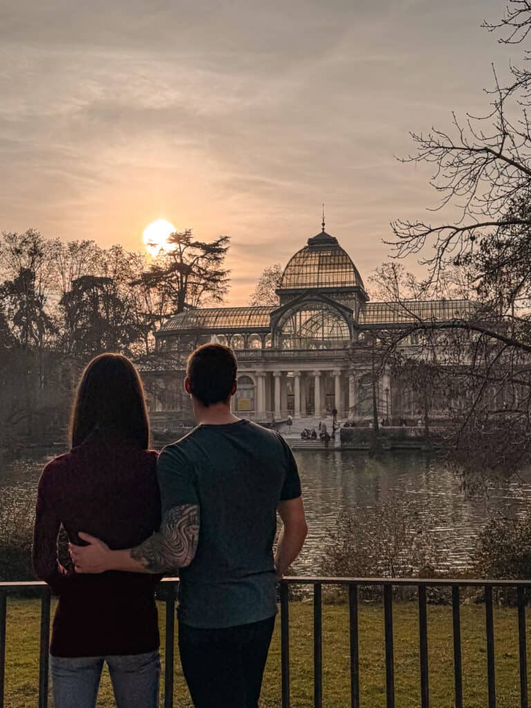 A couple embraces while watching the sunset behind the Crystal Palace in Madrid's Retiro Park, a moment of romance and tranquility by the reflective waters.