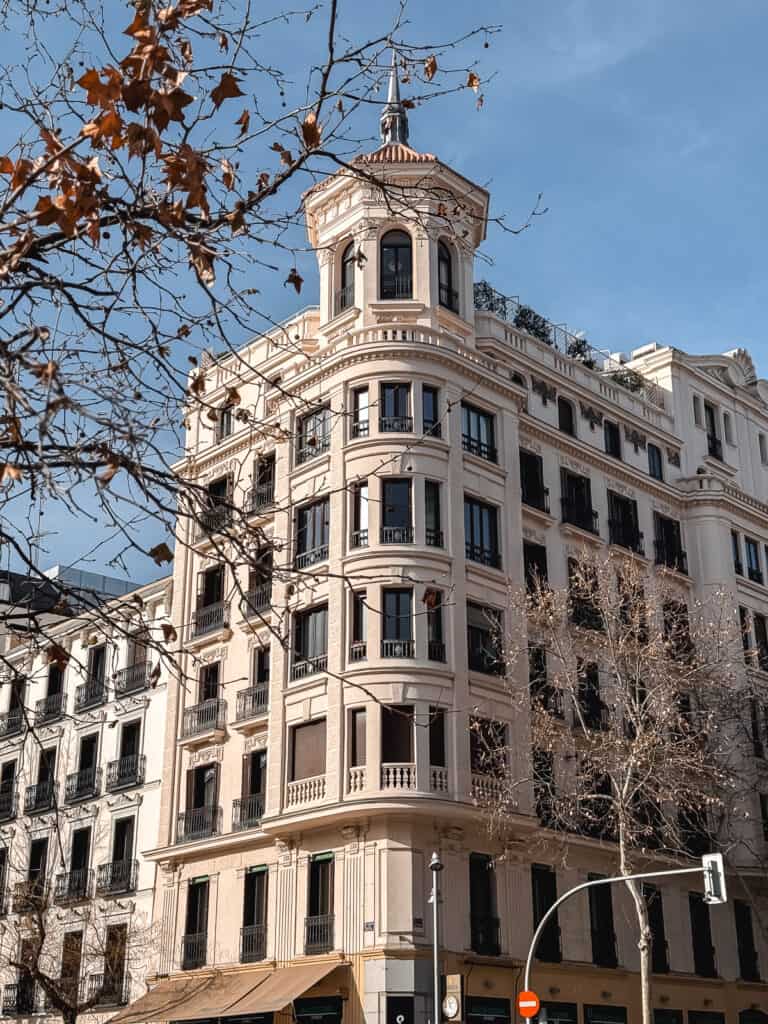 A stately corner building with a round tower and intricate facade stands under a blue sky in Madrid, framed by the bare branches of trees, showcasing the city's historic architecture.