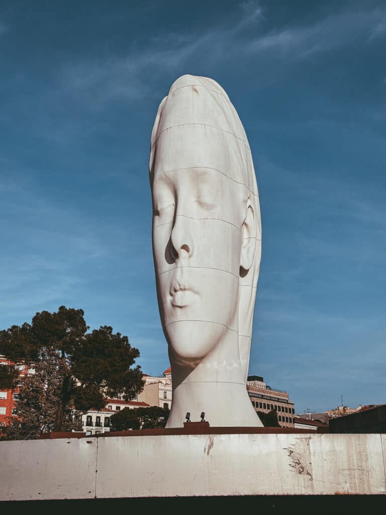 A monumental sculpture of a white, fragmented head stands against a clear blue sky in Madrid, offering an example of modern art in an urban public space.