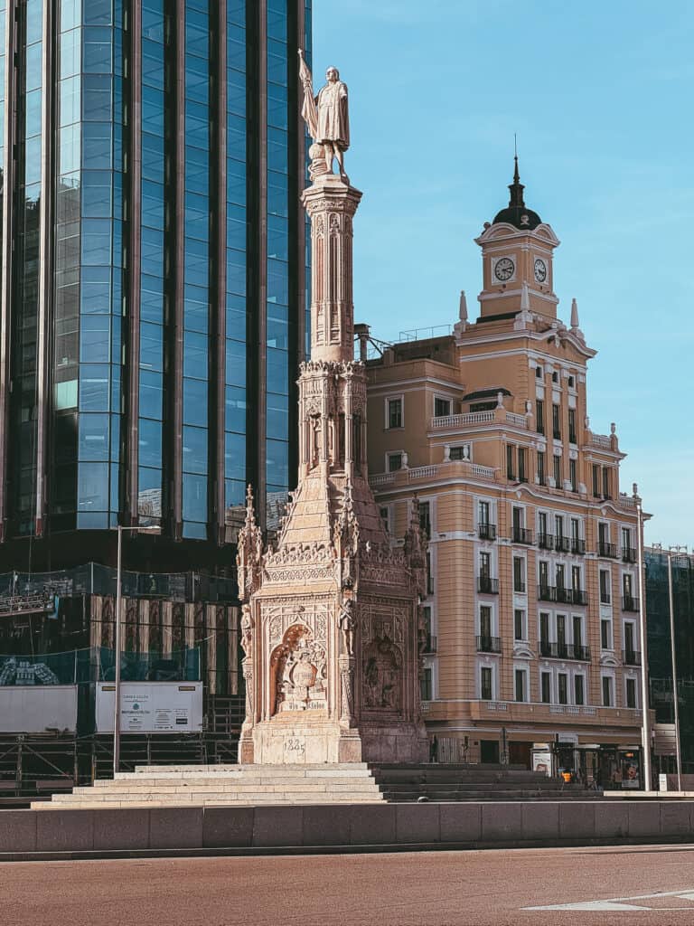 A historical monument featuring a statue atop a decorative column stands in stark contrast to the modern glass building behind it, blending Madrid's rich past with contemporary design.