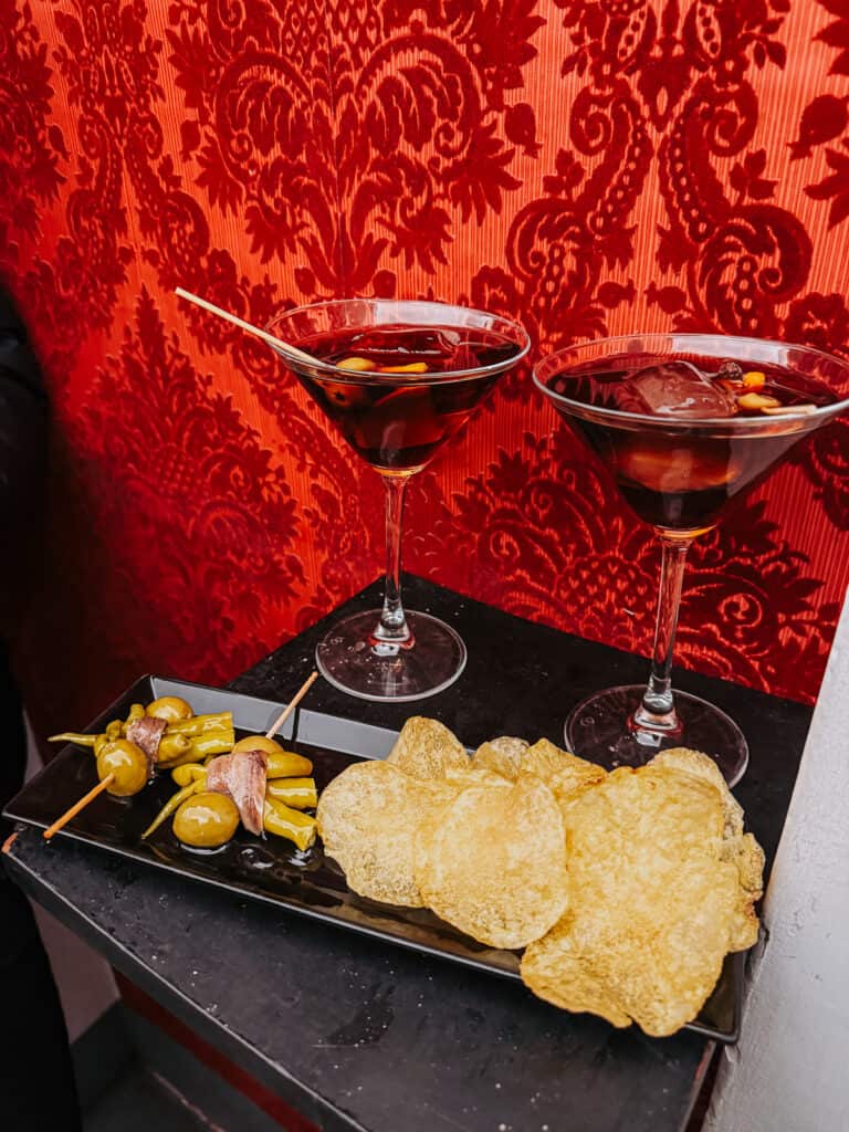 An elegant presentation of traditional Spanish tapas, with vermouth cocktails, potato chips, and skewered olives and anchovies on a red damask-patterned backdrop.