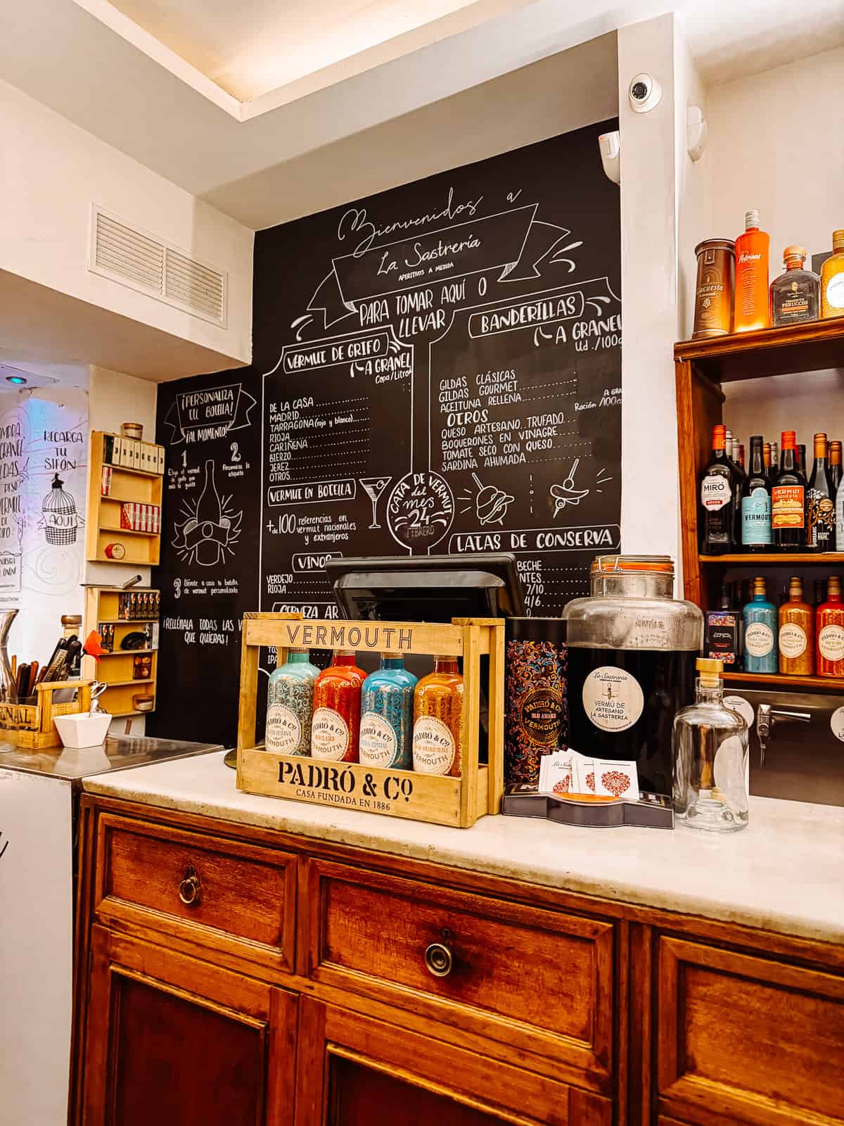 A quaint vermouth bar with a chalkboard menu detailing an array of tapas and drinks, alongside a vintage wooden counter displaying bottles of vermouth, inviting a local Spanish dining experience.
